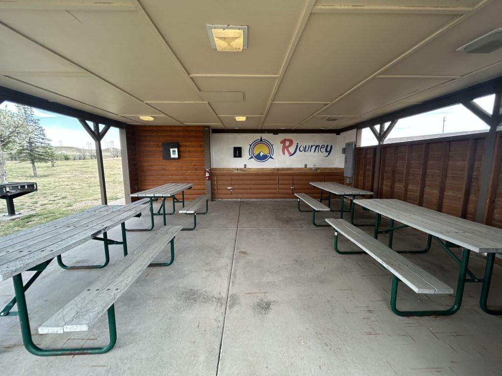 Covered picnic pavilion at Cheyenne RV Resort by RJourney with multiple gray wooden picnic tables on a concrete floor. The back wall features a sink, log siding, and the RJourney logo painted above. A charcoal grill is visible outside on the grassy area.