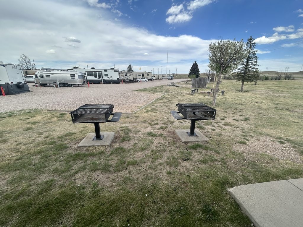 Two metal charcoal grills on concrete pads at Cheyenne RV Resort by RJourney, positioned on a grassy area near the edge of the group tent sites. Several RVs and trailers are parked in the background on a gravel lot under a partly cloudy sky.