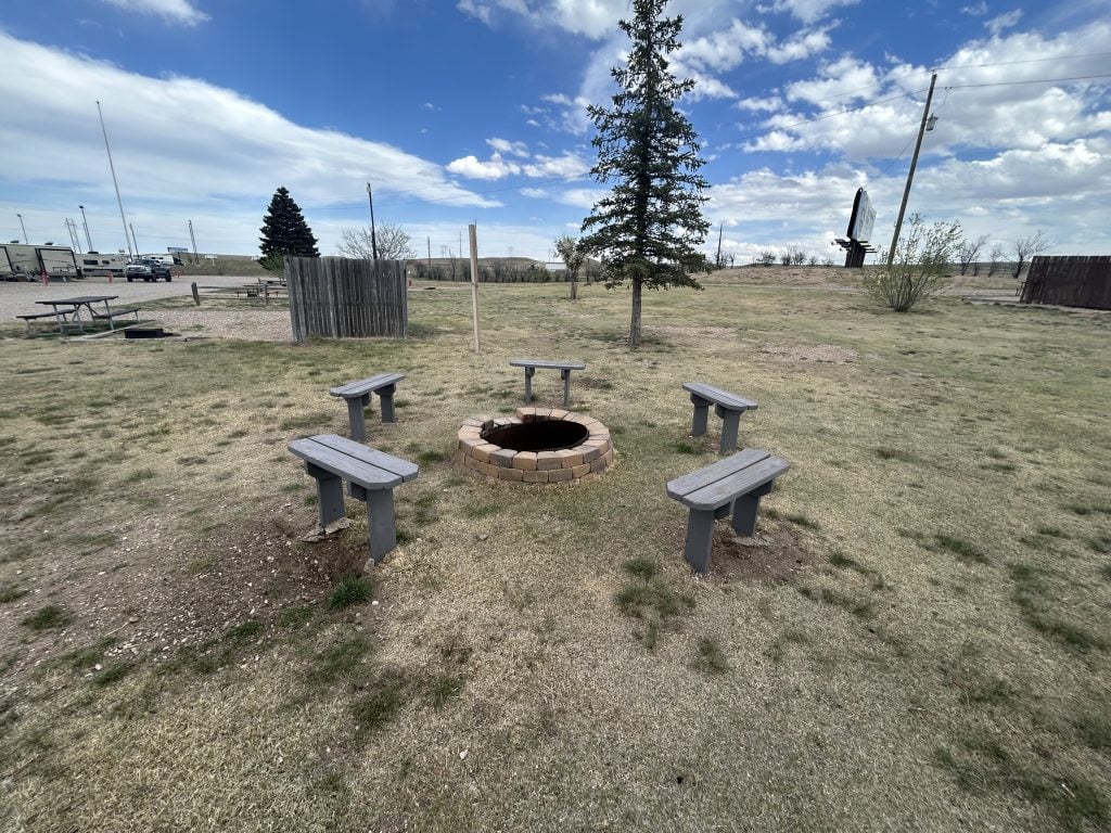 Fire pit area at Cheyenne RV Resort by RJourney featuring a circular stone fire ring surrounded by six wooden benches on a patchy grassy lawn. Picnic tables, fencing, RVs, and a billboard are visible in the background under a partly cloudy sky.