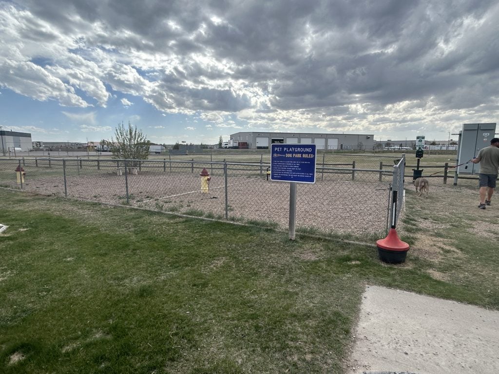 A fenced dog park labeled “Pet Playground” at the Cheyenne RV Resort by RJourney features a gravel play area with decorative fire hydrants, a small tree, and posted park rules, with a man walking a dog near the entrance under a partly cloudy sky.