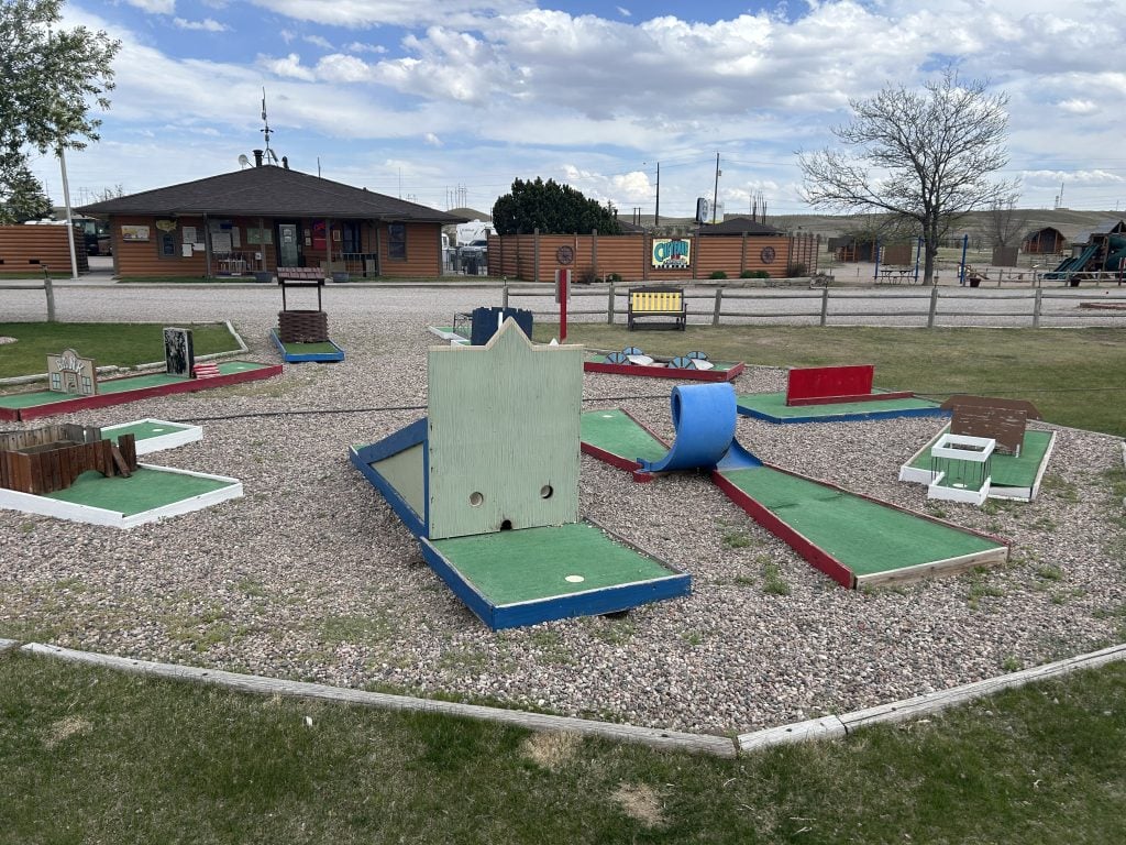 Small outdoor mini golf course at Cheyenne RV Resort by RJourney with various colorful obstacles and turf-lined holes set on a gravel area. The resort’s main office building and nearby playground are visible in the background under a partly cloudy sky.