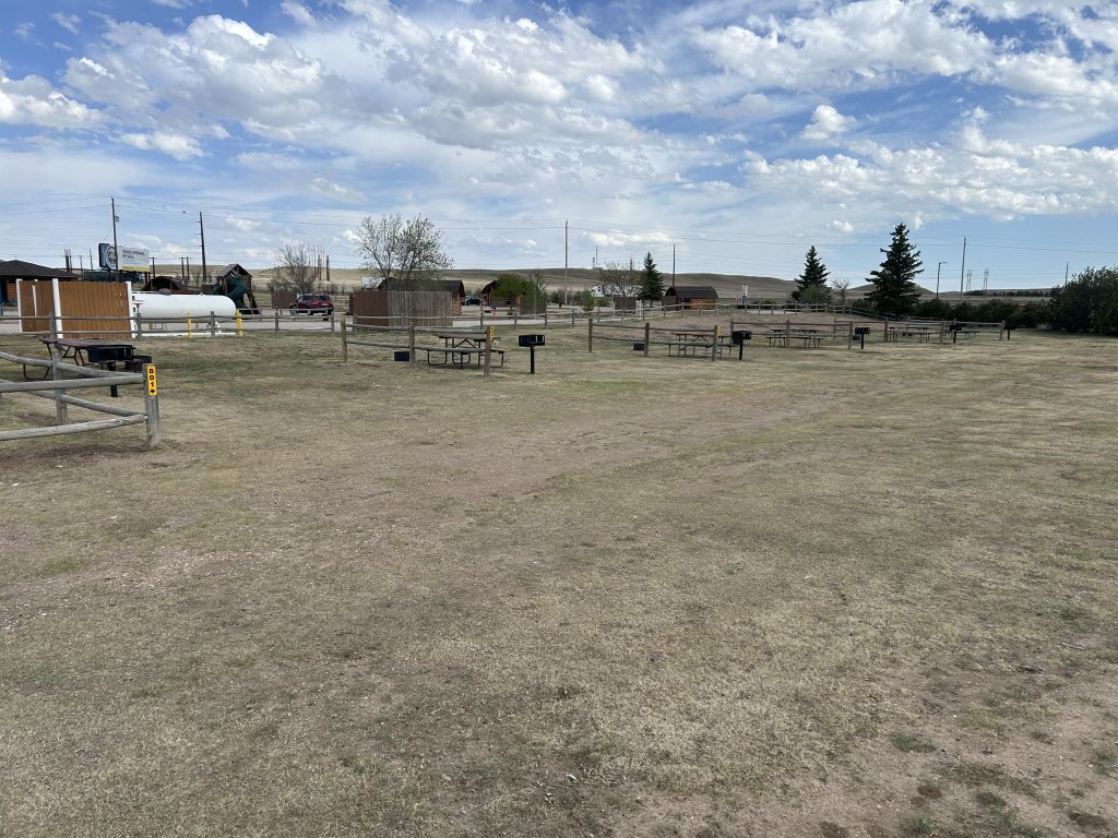 Group tent sites at Cheyenne RV Resort by RJourney featuring multiple picnic tables and charcoal grills spread across a wide, grassy area. The sites are partially divided by wooden fences and privacy panels, with cabins and resort facilities in the background.