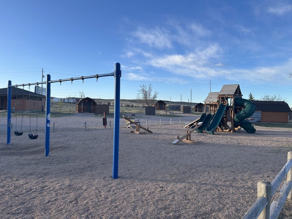 Playground area at Cheyenne RV Resort by RJourney featuring blue swing sets, a large wooden play structure with green slides, and multiple cabins in the background. The playground sits on a gravel surface and is enclosed by a wooden fence under a clear blue sky.