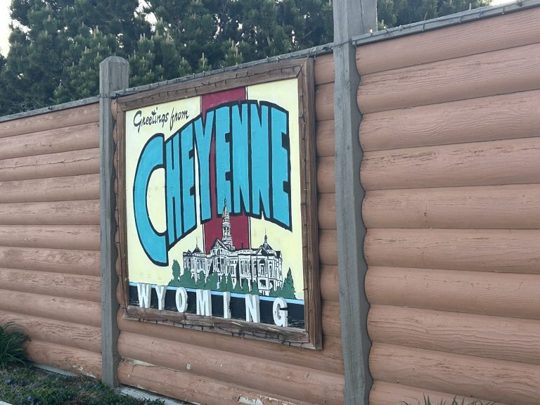 Painted sign reading “Greetings from Cheyenne Wyoming” mounted on a log-style wooden wall at Cheyenne RV Resort by RJourney. The sign features bold turquoise lettering and an illustrated building, likely a local landmark.