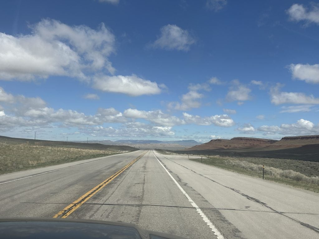 A long, empty highway stretches through open Wyoming landscape with rolling hills and red rock formations in the distance under a bright blue sky with scattered clouds.