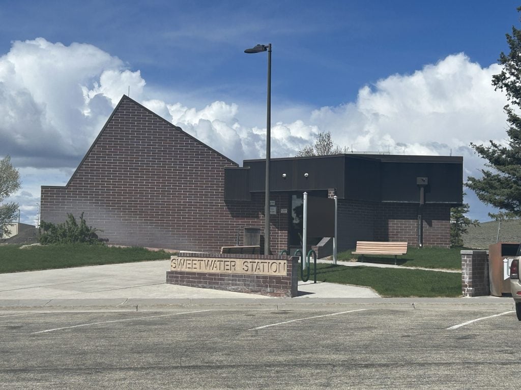 The Sweetwater Station rest area in Wyoming features a modern brick building with a sloped roof, a bench, and a sign out front, set under a bright blue sky with large white clouds.