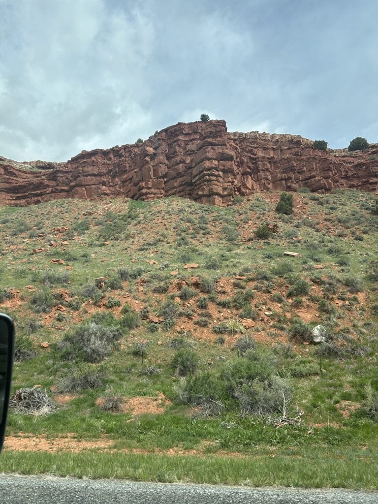 A rugged red rock cliff rises above a grassy, shrub-covered slope beside a road in Wyoming, with scattered small trees on top and a partly cloudy sky overhead.