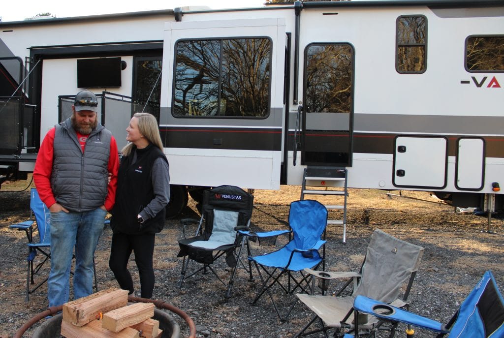 A man (Alan) and woman (Morgan) stand smiling beside a fire pit stacked with wood, in front of a large Valor fifth-wheel RV. Several camping chairs are arranged around the fire, with the RV’s outdoor patio and slide-outs visible behind them.