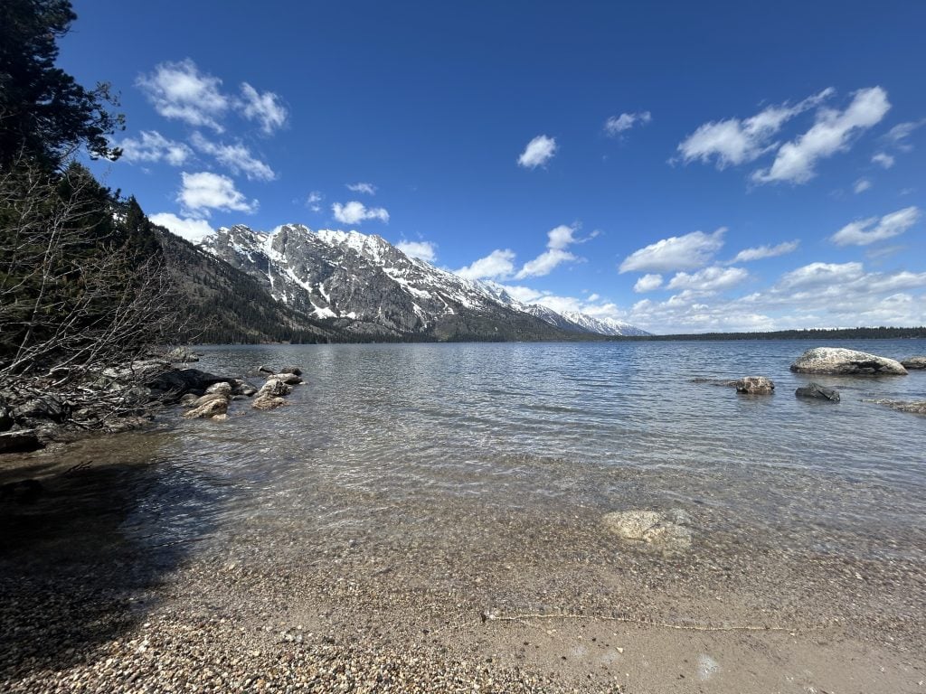 Clear lake water laps against a pebbled shoreline, with large rocks scattered near the edge. Snow-capped mountains rise sharply in the distance under a bright blue sky with scattered clouds.
