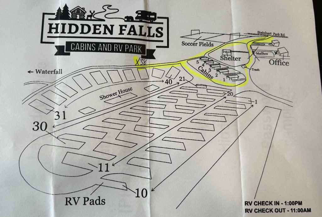 Basic illustrated map of Hidden Falls Cabins & RV Park showing numbered RV pads, cabins, shower house, office, shelter, and soccer fields. A yellow-highlighted path runs through the park with an “X” marking spot 38 near the labeled waterfall. RV check-in time is 1:00 PM and check-out is 11:00 AM.
