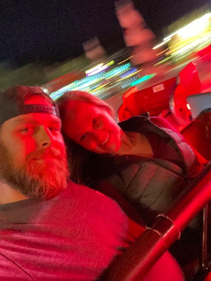 Alan and Morgan smile while riding an amusement park ride at night, illuminated by bright red lighting. The colorful lights of the park blur in the background, capturing the motion and excitement of the moment.
