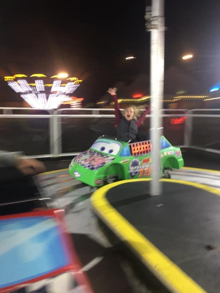 Morgan throws her hands up in excitement while riding a small green race car ride at night. The background is a blur of amusement park lights, emphasizing the motion and energy of the moment.
