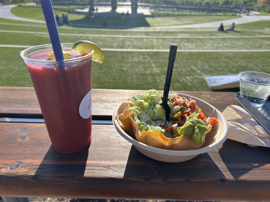 Taco salad in a crispy tortilla bowl topped with lettuce, guacamole, pico de gallo, and jalapeños, served alongside a red frozen drink with a lime wedge from Los Rios Cantina. The meal sits on a wooden table overlooking the green lawn at Cumming City Center.