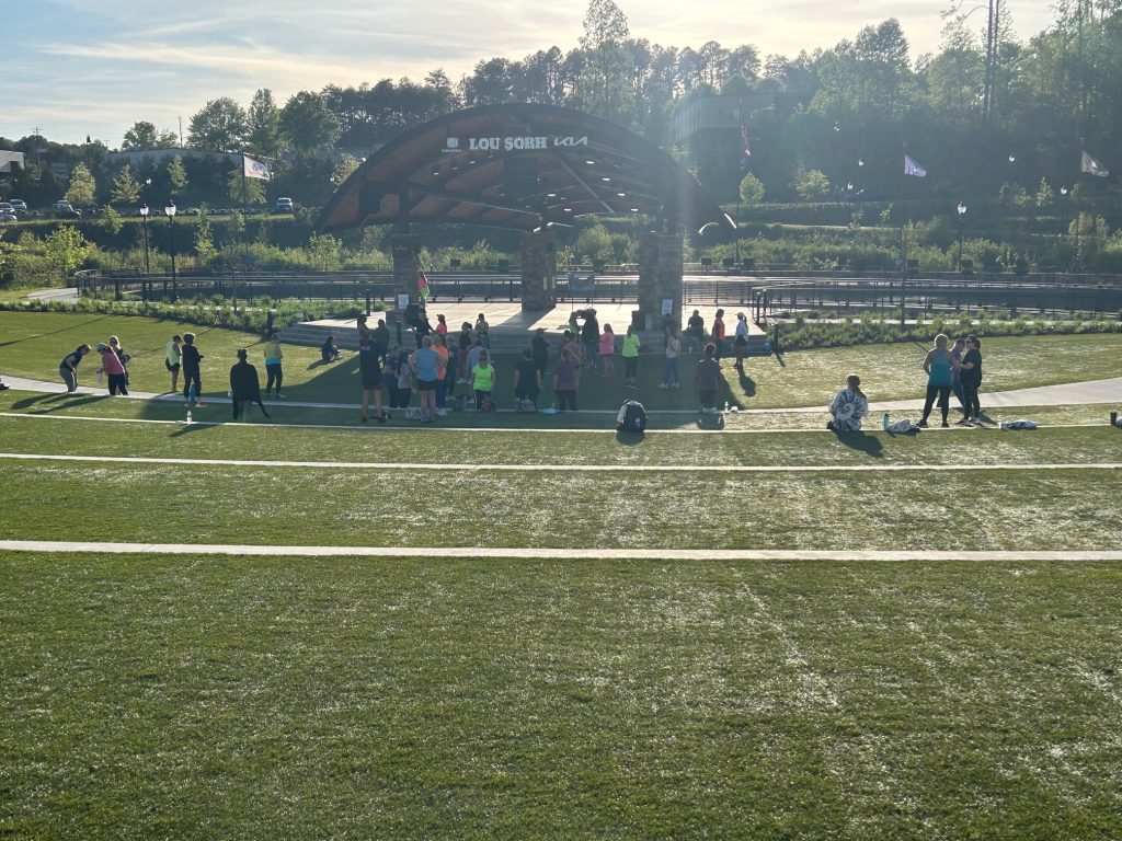 Group of people gathering on a large green lawn in front of the Lou Sobh amphitheater at Cumming City Center before a Zumba class. The scene is lit by late afternoon sun with trees and landscaped walkways in the background.