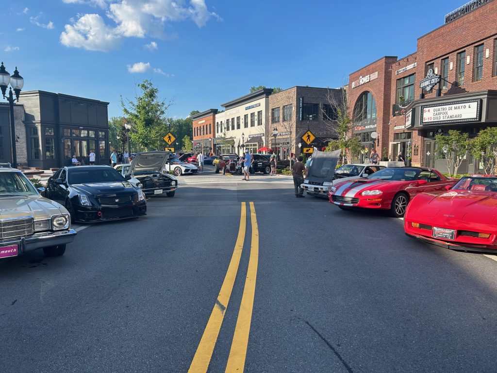 Classic and modern cars lined up on a closed street during a car show at Cumming City Center, with hoods open and people walking between vehicles. Brick buildings, shops, and a movie-style marquee surround the lively event under a bright blue sky.