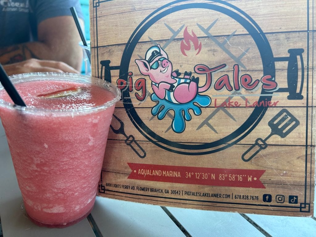 A frozen pink drink with a black straw sits on a table next to a wooden-style menu for Pig Tales Lake Lanier, a waterfront restaurant. The menu features a cartoon pig in sunglasses splashing into blue water with fire and grilling tools in the background, along with location coordinates and contact info.
