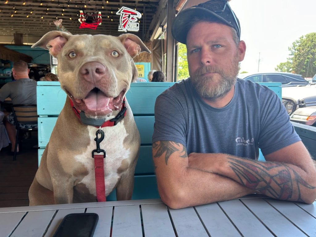 A happy, tan and white dog with a red leash sits next to a bearded man with crossed arms and tattooed forearms at an outdoor restaurant. They're seated at a turquoise booth under string lights, with neon beer signs and other diners in the background.