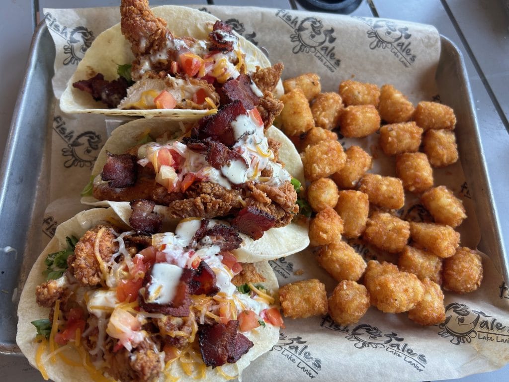 Three flour tortillas filled with crispy fried chicken, diced tomatoes, shredded cheese, bacon pieces, and drizzled ranch dressing are served on branded paper. A generous side of golden tater tots fills the other half of the tray.