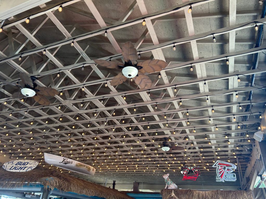 Ceiling of a waterside-themed restaurant decorated with string lights, wooden beams, and palm leaf ceiling fans. Hanging decor includes upside-down canoes branded with Bud Light and Louie’s Summer Shandy, along with neon signs for Budweiser and the Atlanta Falcons.