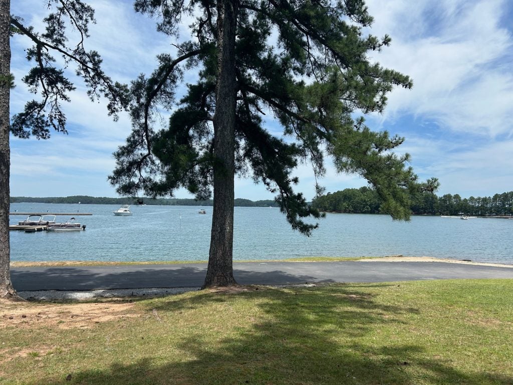 Peaceful lakeside view at Lake Lanier featuring calm blue water, several boats, and a dock to the left. Tall pine trees frame the scene with a grassy shoreline and a paved path in the foreground under a partly cloudy sky.