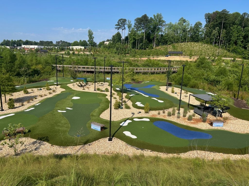 Brightly colored mini golf course at Cumming City Center featuring artificial turf greens, blue water features, white sand traps, and landscaping with gravel and shrubs. Surrounded by trees and a wooden pedestrian bridge in the background under a clear blue sky.