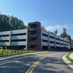Multi-level parking deck at Cumming City Center, with concrete and brick exterior, surrounded by trees and landscaped sidewalks under a clear blue sky. A vertical sign on the stair tower reads "PARK" and cars are visible on the ground level.