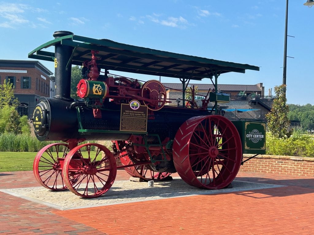 Restored steam tractor on display at Cumming City Center, featuring red wheels, black and green body, and historical plaques. The vintage engine sits on a brick plaza with shops and greenery in the background under a clear blue sky.
