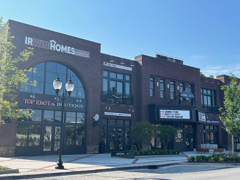 Brick building at Cumming City Center featuring multiple businesses including Top Knotch Boutique, Unique Cuts Bar, and Lilly Rose Co. Marketplace. A black marquee sign displays upcoming events, and the scene is framed by trees, street lamps, and a clear blue sky.