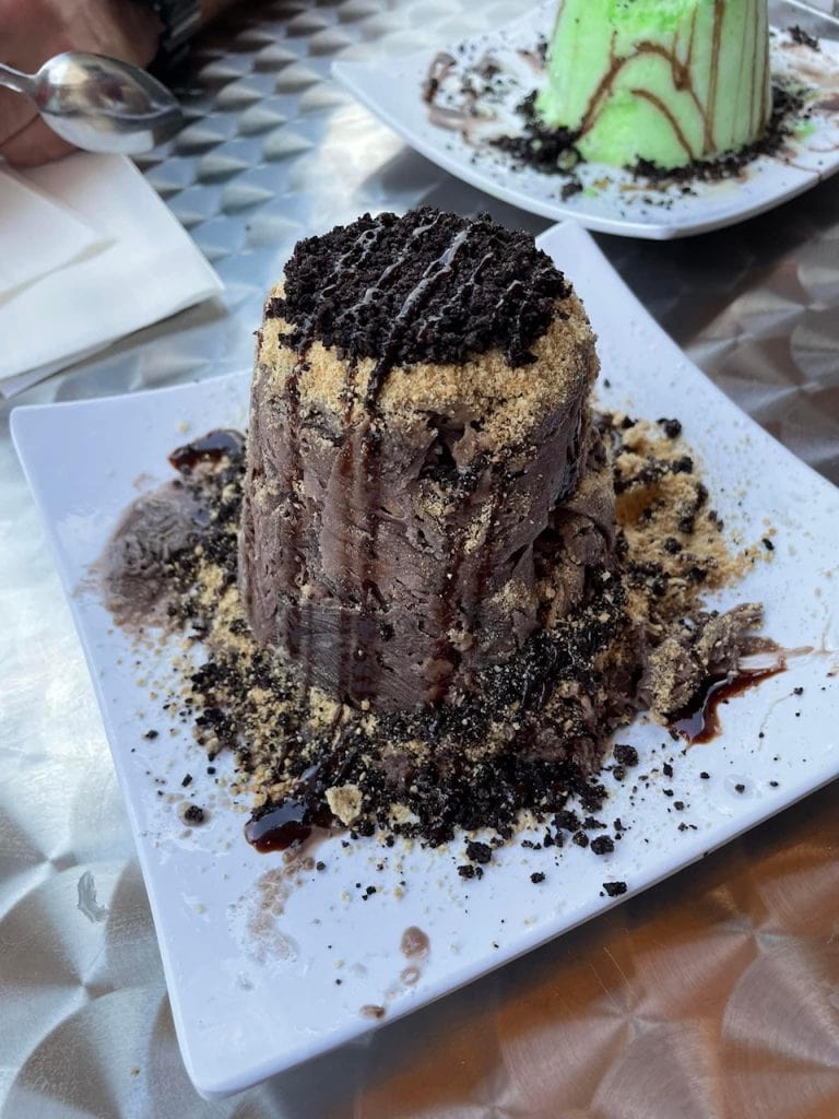 Chocolate shaved snow dessert from Vampire Penguin, topped with crushed Oreos and graham cracker crumbs, and drizzled with chocolate syrup. Served on a white plate with cookie crumble scattered around the base.