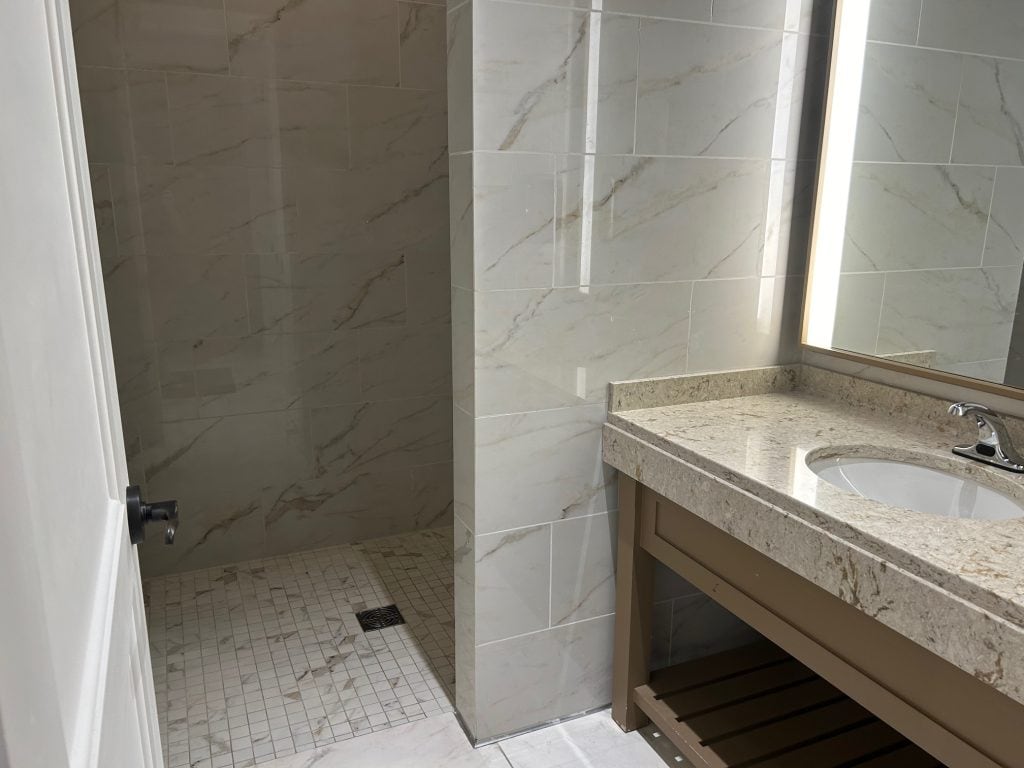 A modern bathroom features a walk-in shower with beige marble-patterned tile and a square drain. Beside it, a granite countertop with an under-mount sink and illuminated mirror adds a sleek, clean look.