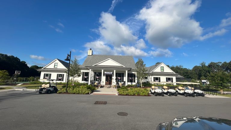 Front view of the clubhouse at Hilton Head National RV Resort on a sunny day, featuring white siding, black shutters, and patriotic bunting on the windows. Several golf carts are parked out front, and a basketball hoop is visible to the left near landscaped greenery.
