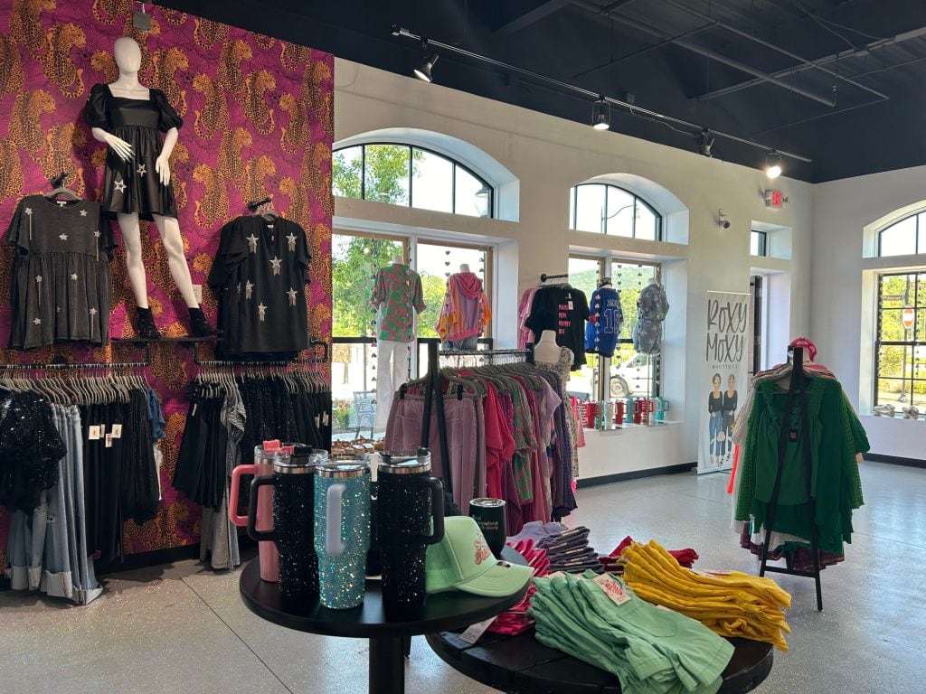 Interior of Roxy Moxy Boutique at Cumming City Center showcasing colorful women’s clothing, glitter tumblers, and accessories. A bold pink and leopard print wall features star-accented outfits, while racks display a mix of casual and statement pieces under bright natural light.