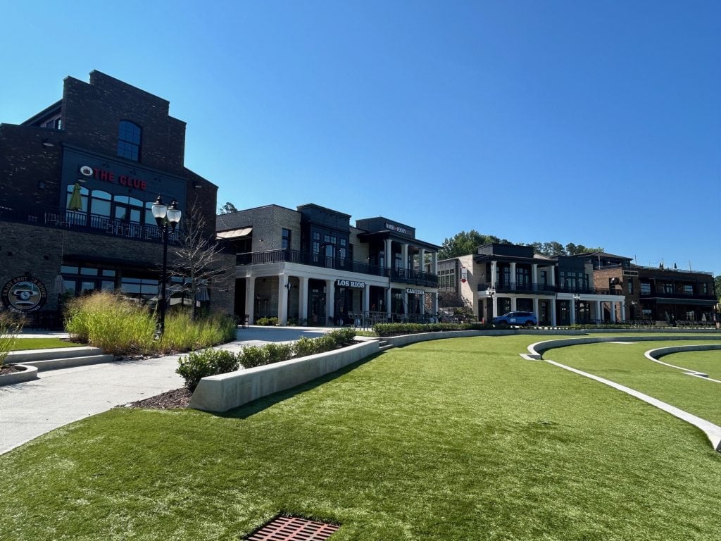 Row of modern brick and stone buildings at Cumming City Center, including The Club and Los Rios Cantina, viewed from the edge of a curved green lawn amphitheater. Clear blue sky and landscaped walkways enhance the inviting, open-air setting.
