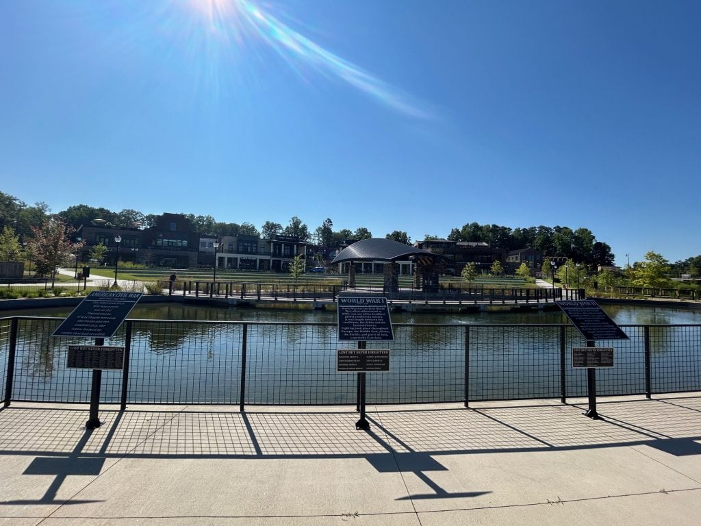 View across a calm pond at Cumming City Center with memorial plaques in the foreground honoring fallen soldiers from American wars. Shops and buildings line the opposite side of the water, and the scene is lit by bright morning sun under a clear blue sky.