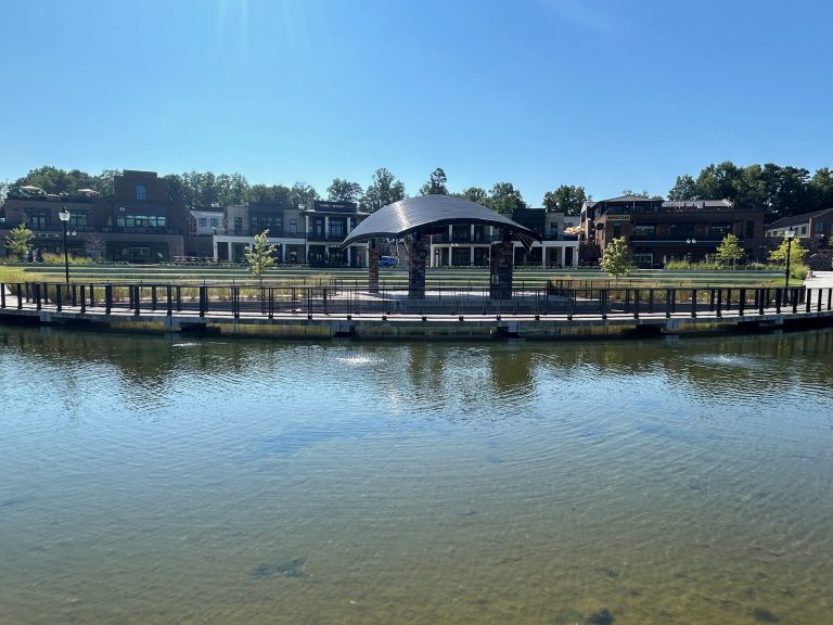 Scenic view of a covered pavilion on a boardwalk surrounded by a pond at Cumming City Center, with reflections in the water and a backdrop of shops and restaurants. The sky is clear and blue, highlighting the calm and inviting setting.
