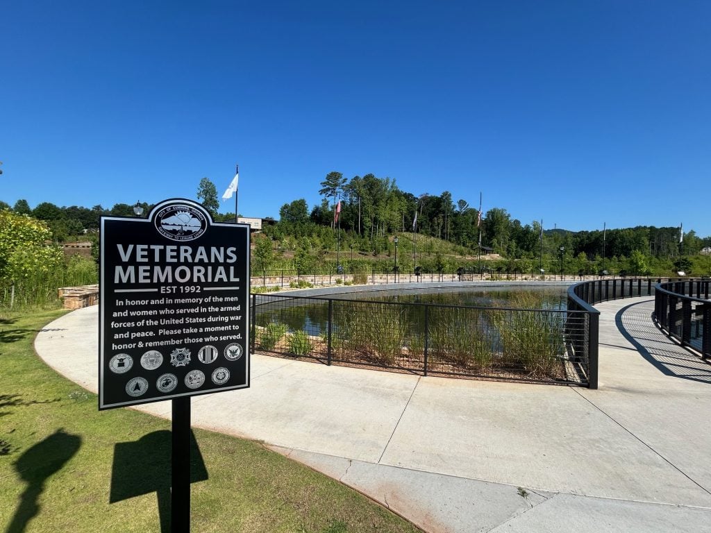 Black and white sign for the Veterans Memorial at Cumming City Center stands beside a curved walkway overlooking a landscaped pond. The sign honors U.S. armed forces members and is set against a backdrop of trees, flagpoles, and a clear blue sky.