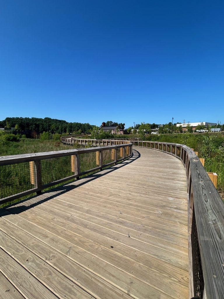 Curved wooden pedestrian bridge at Cumming City Center with mesh railings, leading toward trees and distant buildings under a clear blue sky. The bridge is elevated over green vegetation and casts long shadows on its planks.