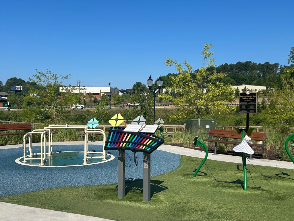 Colorful and accessible play area at Cumming City Center featuring musical instruments, a wheelchair-friendly merry-go-round, and flower-shaped structures. Benches and a sign for the Cumming Kiwanis Club Children’s Park are visible in a landscaped setting under a clear blue sky.