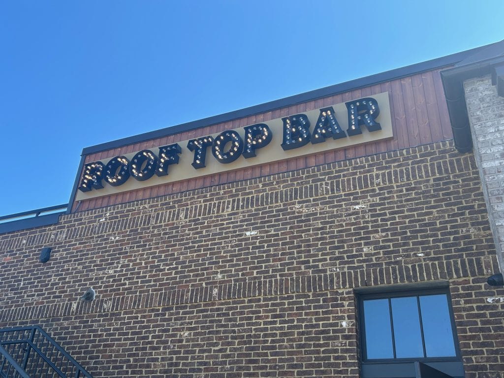 Sign reading “Roof Top Bar” in bold, marquee-style letters with lights, mounted on a brick building at Cumming City Center. The photo is taken from below against a bright blue sky.