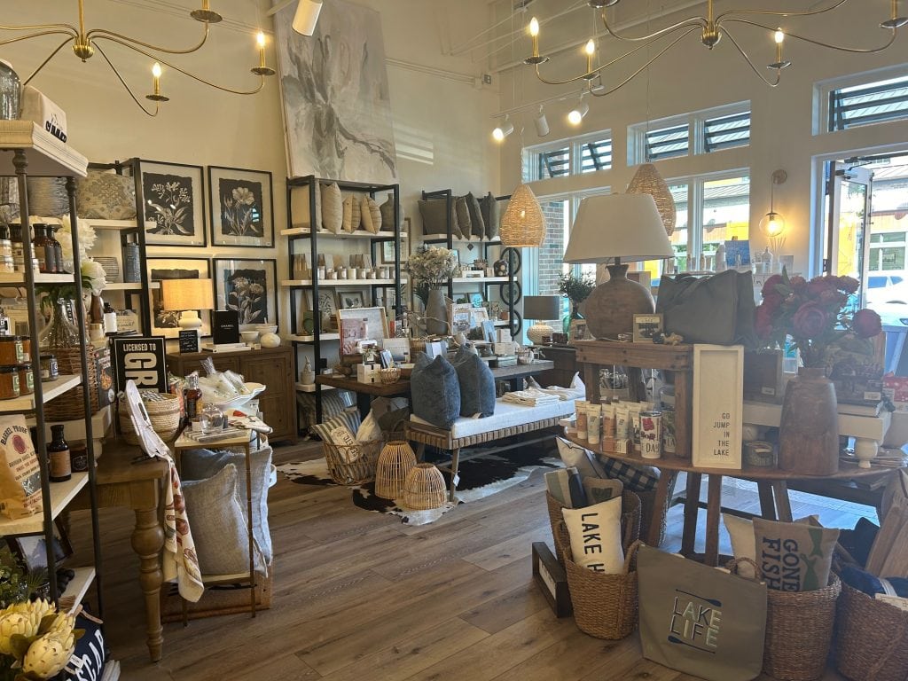 Cozy boutique shop at Cumming City Center featuring home décor, pillows, lamps, framed art, and lake-themed gifts arranged on wooden tables and shelves. Warm lighting and natural textures create an inviting atmosphere with a rustic-modern style.
