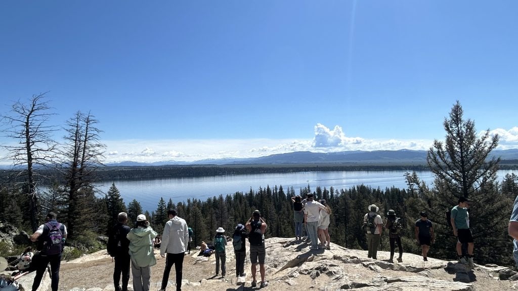 Hikers stand at Inspiration Point overlooking Jenny Lake, with clear blue skies and distant mountain ranges enhancing the panoramic view. The rocky overlook is surrounded by pine trees and offers a scenic reward at the end of the Jenny Lake hike to Inspiration Point.