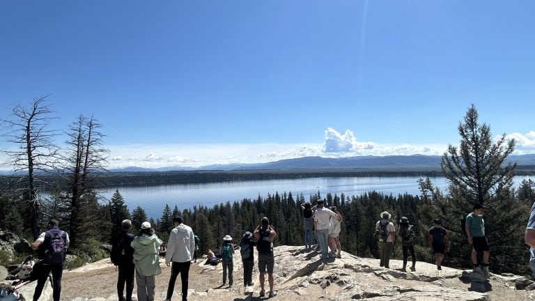 Hikers stand at Inspiration Point overlooking Jenny Lake, with clear blue skies and distant mountain ranges enhancing the panoramic view. The rocky overlook is surrounded by pine trees and offers a scenic reward at the end of the Jenny Lake hike to Inspiration Point.