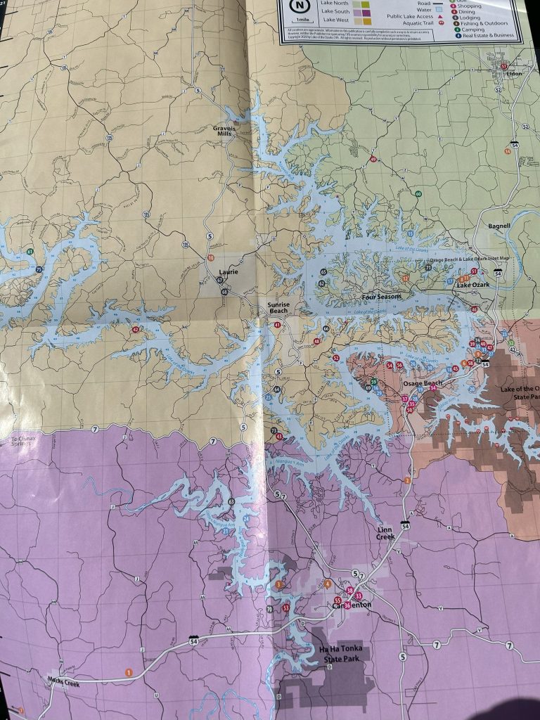 A detailed map of the Lake of the Ozarks region in Missouri, showing roads, towns, lake arms, and highlighted locations for shopping, dining, lodging, camping, and outdoor activities, with color-coded regions and a legend in the top right corner.