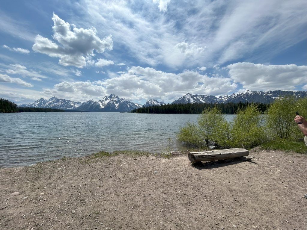 Snow-capped mountains rise behind a calm lake along the Lakeshore Trail in Grand Teton National Park. A log bench rests near the shoreline with sparse greenery and partly cloudy skies above, highlighting the serene setting of this easy hike.
