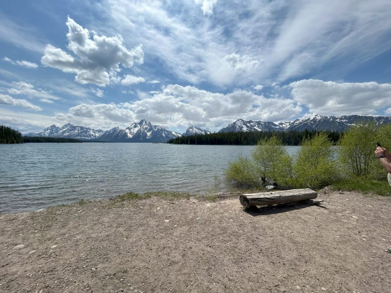Snow-capped mountains rise behind a calm lake along the Lakeshore Trail in Grand Teton National Park. A log bench rests near the shoreline with sparse greenery and partly cloudy skies above, highlighting the serene setting of this easy hike.