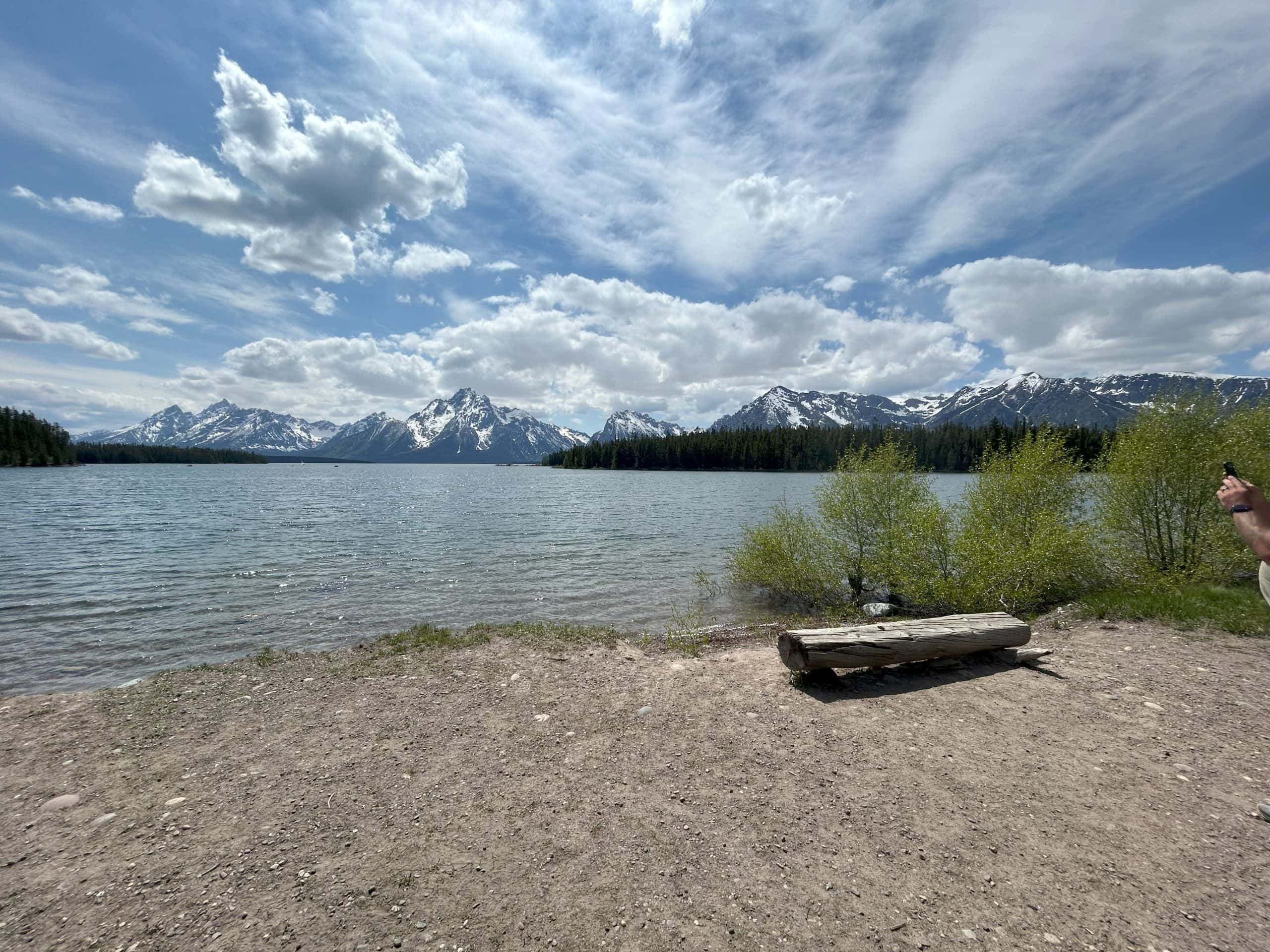 Snow-capped mountains rise behind a calm lake along the Lakeshore Trail in Grand Teton National Park. A log bench rests near the shoreline with sparse greenery and partly cloudy skies above, highlighting the serene setting of this easy hike.