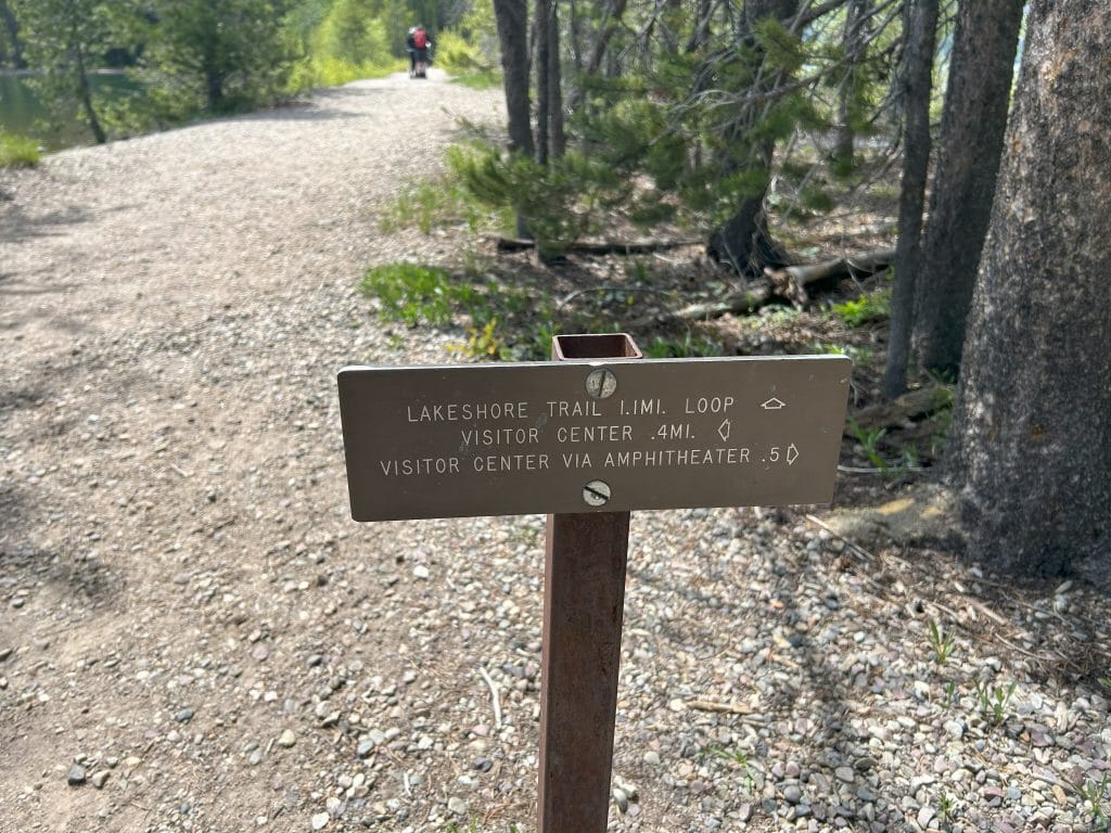 Trail signpost along the Lakeshore Trail in Grand Teton National Park, showing directions for a 1.1-mile loop and routes to the visitor center, including a 0.4-mile path and a 0.5-mile option via the amphitheater. The easy hike path is shaded by trees with a gravel trail leading into the distance.