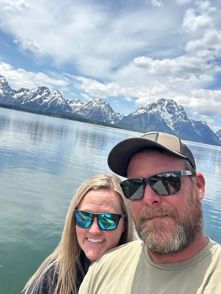 A couple poses for a selfie in front of the calm lake and snow-covered peaks of the Teton Range along the Lakeshore Trail in Grand Teton National Park. Their sunglasses reflect the scenic view, capturing the beauty of this easy hike and peaceful lakeside moment.