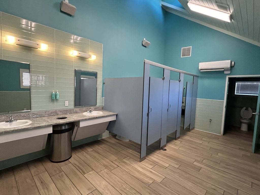 Clean and brightly lit bathhouse with turquoise walls, light wood-look tile flooring, and multiple toilet stalls. A granite countertop with two sinks and wall-mounted soap dispensers sits beneath large mirrors.