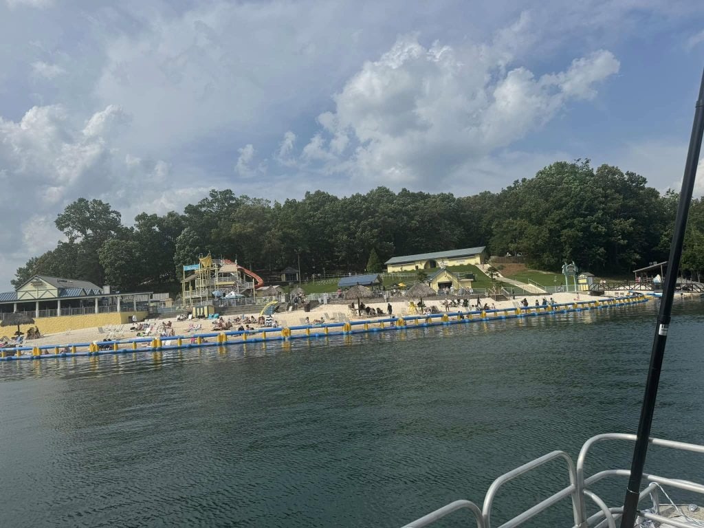 View from the water of a sandy beach at Margaritaville, with people lounging near the shore and a floating inflatable barrier in the lake. Behind the beach, colorful water slides, buildings, and lush green trees create a lively resort atmosphere.